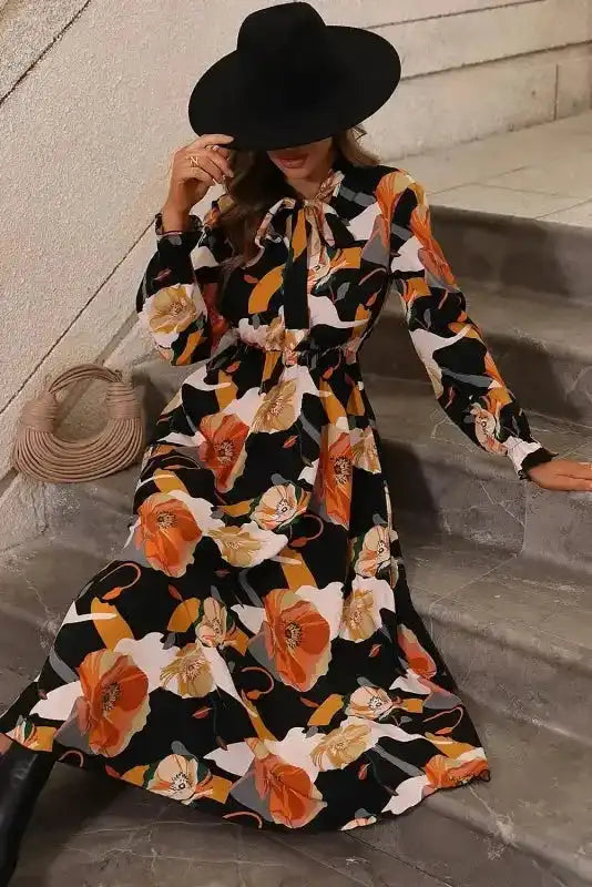 Model seated in orange floral long sleeve dress with tie neck, showcasing soft fabric and bold floral print for casual wear.