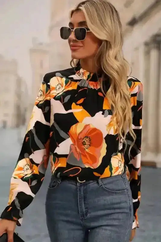 Model in orange floral blouse with long sleeves and ruffled neck, styled with jeans and sunglasses for a trendy look.