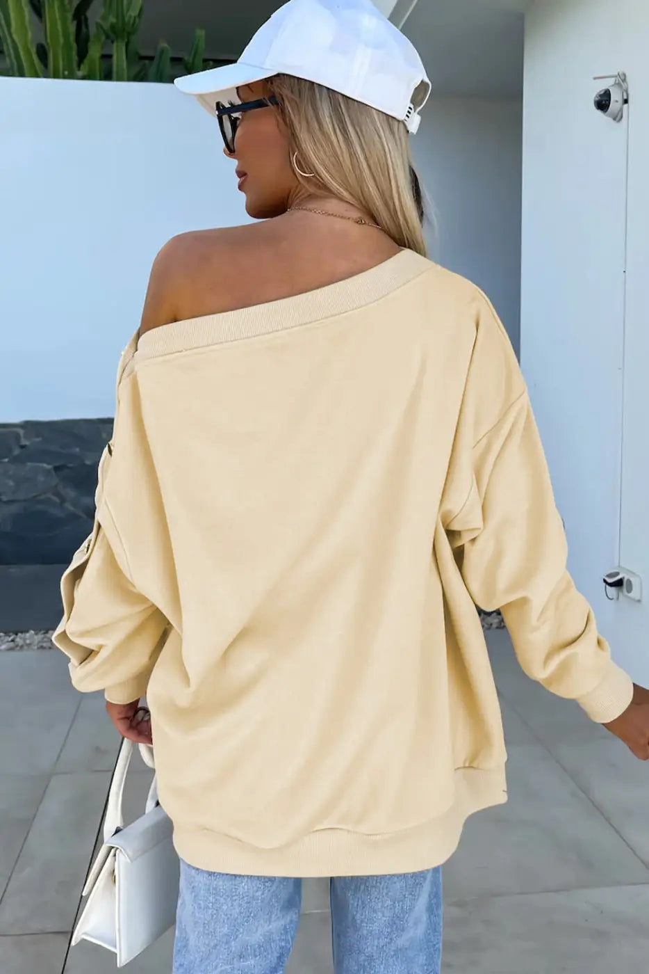 Off-shoulder pale yellow relaxed fit sweater worn with a white baseball cap and blue jeans
