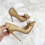 Handheld view of beige Riviera Bow Court Heels with spiked trim and bow, highlighting their stylish stiletto silhouette.