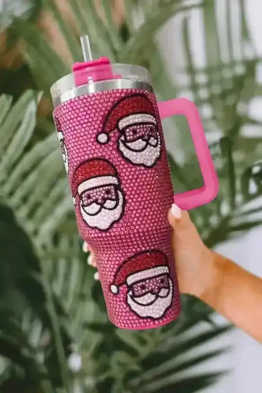 Playful Santa Claus thermos cup with sparkling rhinestones and pink straw lid, held against tropical green background, keeps drinks hot or cold.
