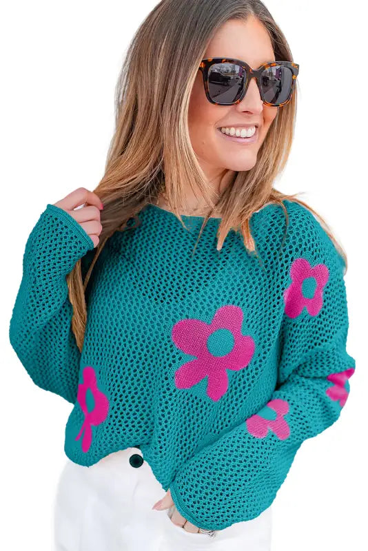 A smiling woman in sunglasses wears a hollowed knit teal mesh sweater with pink flower designs