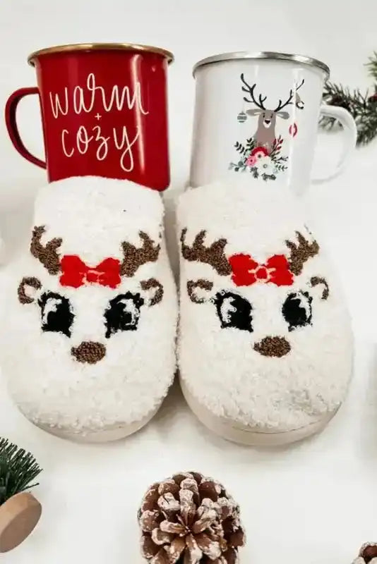 White plush reindeer slippers with red bows displayed with holiday mugs, perfect gift idea for cozy Christmas lounging.