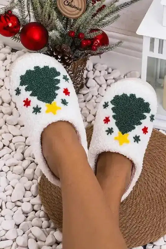 Feet in white plush slippers featuring Christmas tree and star embroidery, cozy holiday slippers for indoor comfort.