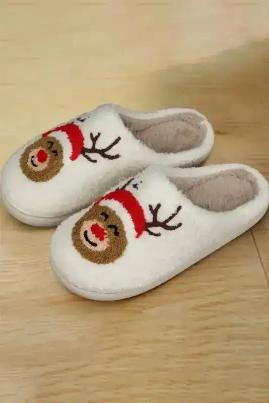 Pair of white Christmas deer plush slippers with reindeer face and red Santa hat, displayed on wooden floor for holiday apparel.