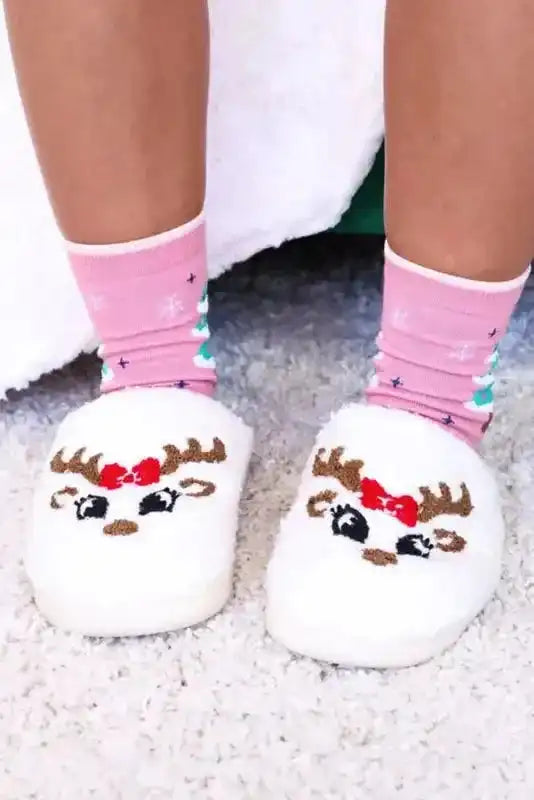 Feet in white Christmas deer slippers with red bows and antlers, paired with festive socks for holiday comfort.